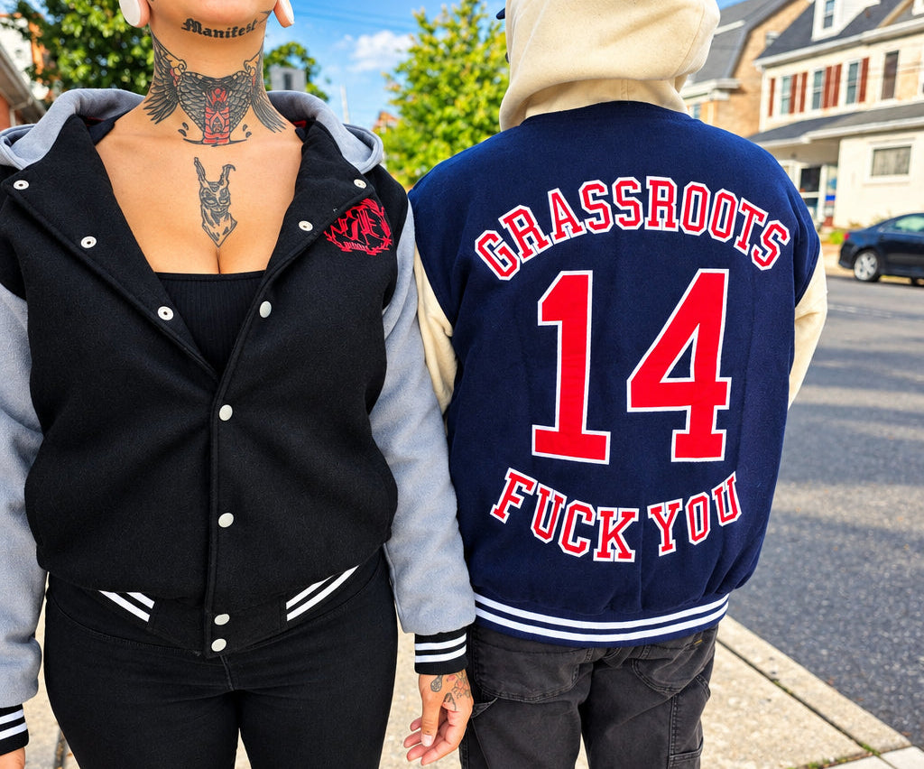 Two people standing outdoors on a sidewalk. The person on the left wears a black varsity-style jacket with gray sleeves and a red embroidered patch.